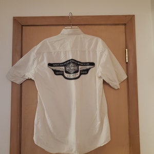 Op de afbeelding: Een wit overhemd met korte mouwen en knopen met een zwart Harley-Davidson-logo op de rug. Het logo bevat de tekst "Ninety-Five Years of Great Motorcycles" en de jaren "1903 1998 2003".