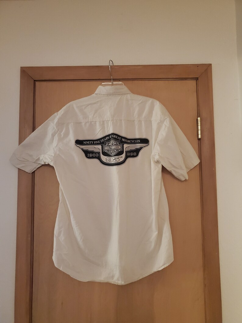 Op de afbeelding: Een wit overhemd met korte mouwen en knopen met een zwart Harley-Davidson-logo op de rug. Het logo bevat de tekst "Ninety-Five Years of Great Motorcycles" en de jaren "1903 1998 2003".