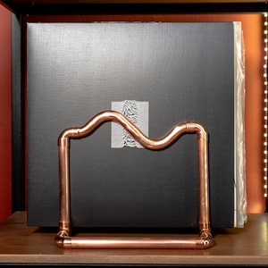 May include: A black vinyl record album with a copper pipe record stand. The album cover has a white graphic of a wave pattern.