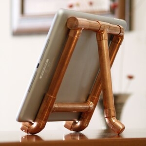 May include: A copper pipe tablet stand with a silver tablet resting on top. The stand is made of copper pipes that are connected together to form a triangular shape. The stand is sitting on a wooden surface.