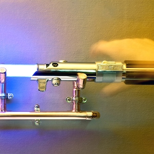 May include: A silver and black lightsaber with a blue and white glowing blade, mounted on a copper-colored metal stand. The lightsaber is held in place by two brackets. The background is a gradient of purple and yellow.