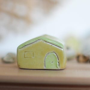 Op de afbeelding: Een kleine keramische huisfiguur met een gele behuizing en een groen dak. Het huis heeft een kleine deur en een raam en staat op een houten oppervlak. Het beeldje is een decoratief item.