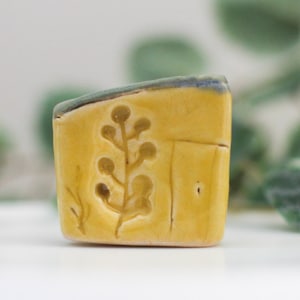 May include: A small, rectangular ceramic piece with a yellow base and a carved botanical design. The top edge is a muted green with a thin blue stripe. The design features a stylized plant with rounded leaves.