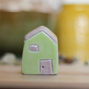 Op de afbeelding: Een klein, handgemaakt keramisch huisje met een lichtgroen lichaam en een lavendelkleurig dak, deur en raam. Het huisje is een miniatuur decoratie-artikel, met een glanzende afwerking. Een gele pot is wazig op de achtergrond.