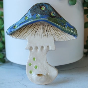 May include: A ceramic mushroom figurine with a blue and green cap and a cream-colored stem. The cap has a textured surface with small green dots. The mushroom is set against a white background with green foliage.