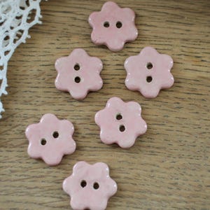 May include: Six light pink, flower-shaped buttons with two holes each are arranged on a wooden surface. A white lace trim is visible in the upper left corner. The buttons have a glossy finish.