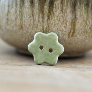 Könnte beinhalten: Ein hellgrüner, blütenförmiger Knopf mit zwei Löchern. Der Knopf ist aus Keramik und hat eine glatte, glänzende Oberfläche. Der Knopf befindet sich vor einem verschwommenen Hintergrund eines Keramiktopfes.