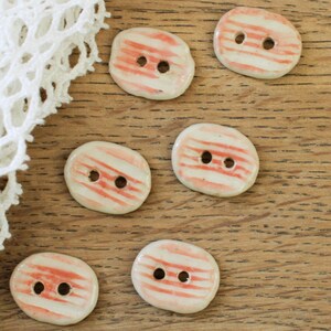 May include: Six ceramic buttons with a cream base and red striped pattern. Each button has two holes for sewing. The buttons are displayed on a wooden surface, with white lace trim in the upper left corner.