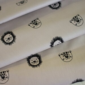 May include: Off-white fabric with a repeating pattern of black animal faces, including lions, leopards, and hedgehogs. The fabric is folded, showing the texture and design. The illustrations are simple and cartoon-like, creating a playful aesthetic.
