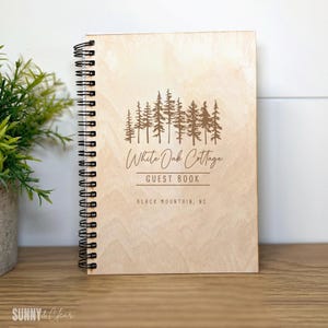 May include: A light-colored wooden guest book with a spiral binding. The cover features a forest silhouette and the text "White Oak Cottage Guest Book, Black Mountain, NC."