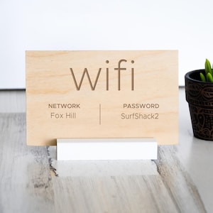 May include: A wooden WiFi sign with engraved text. The word "wifi" is at the top, with "NETWORK" and "PASSWORD" below. The network name is "Fox Hill" and the password is "SurfShack2". The sign is on a white stand.