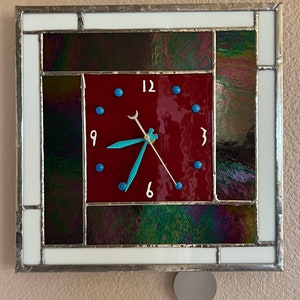 May include: A square stained glass clock with a red face and black, green, and purple accents. The clock face has blue dots around the edge and white numbers for 12, 3, 6, and 9. The clock hands are white and turquoise.