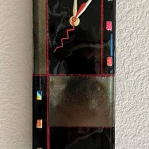 Op de afbeelding: Een zwarte en grijze glazen wandklok met een rood en wit wijzerplaat. De klok is ingelijst in een zwart en grijs geometrisch patroon met kleurrijke accenten.