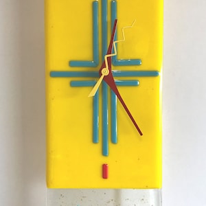 May include: A rectangular wall clock with a bright yellow face. The clock features turquoise and red accents, including a cross-shaped design and a red clock hand. The bottom section is clear with decorative elements.