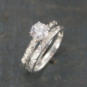 May include: A silver engagement ring set with a large, clear gemstone and a matching band. Both rings feature an intricate, engraved floral pattern. The rings are displayed on a dark, textured surface, showcasing their reflective qualities.