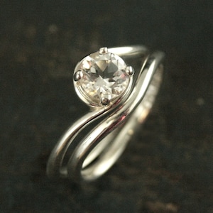 May include: A silver ring set with a clear round gemstone. The band is a wavy design with a second band that wraps around the top of the gemstone.