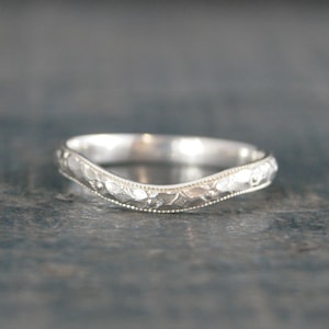 Könnte beinhalten: Ein silberfarbener Ring mit geschwungenem Design. Der Ring hat ein aufwendiges Blumenmuster entlang des Bandes, mit einem zarten, perlenbesetzten Rand. Der Ring ist wahrscheinlich aus Sterlingsilber oder einem ähnlichen Metall gefertigt.