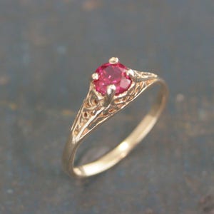 May include: A gold-colored ring featuring a vibrant, round, red gemstone. The ring has an ornate, filigree design on the band, adding a vintage or antique aesthetic. The gemstone is the focal point, set in a four-prong setting.