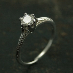 K&ouml;nnte beinhalten: Ein silberner Verlobungsring mit einem einzelnen runden Diamanten, der in einer Krappenfassung sitzt. Die Ringschiene ist mit einem zarten floralen Design verziert.