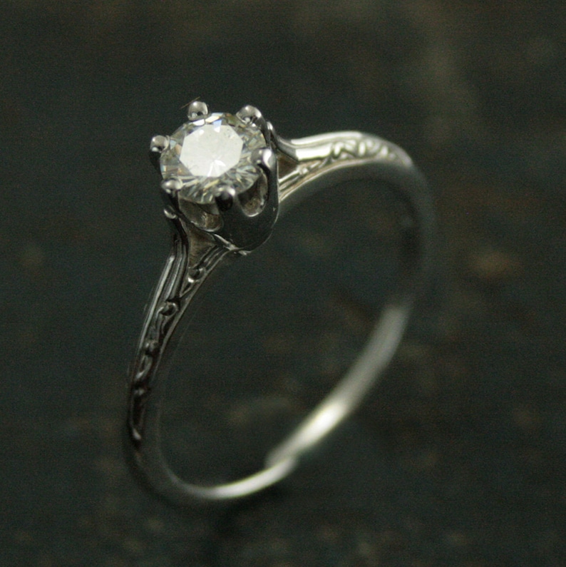K&ouml;nnte beinhalten: Ein silberner Verlobungsring mit einem einzelnen runden Diamanten, der in einer Krappenfassung sitzt. Die Ringschiene ist mit einem zarten floralen Design verziert.