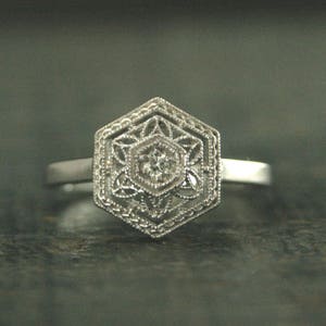 May include: A silver ring with a hexagonal design featuring a delicate filigree pattern and a small, clear gemstone in the center.