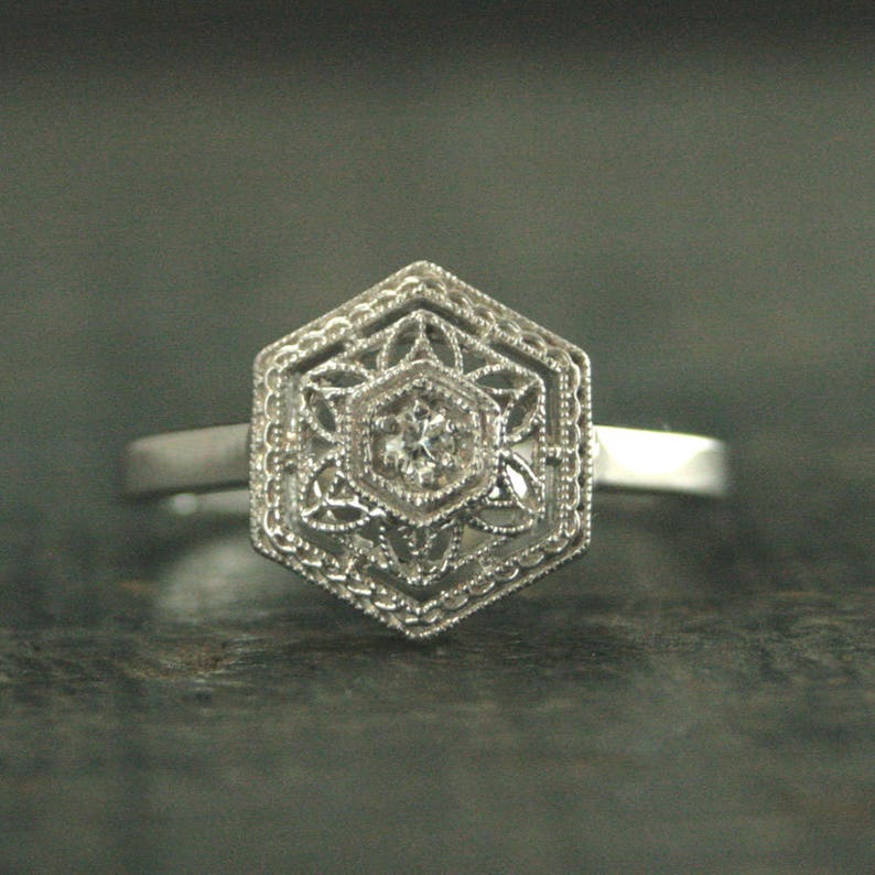 May include: A silver ring with a hexagonal design featuring a delicate filigree pattern and a small, clear gemstone in the center.