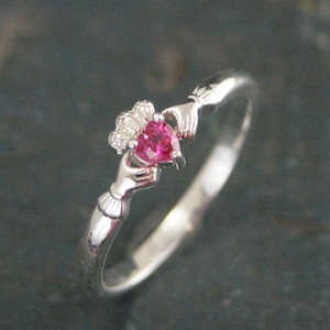 Peut inclure: Bague Claddagh en argent avec une pierre précieuse rose vif en forme de cœur. Le cœur est tenu par deux mains et une couronne est placée au-dessus du cœur. La bague est sur fond sombre.