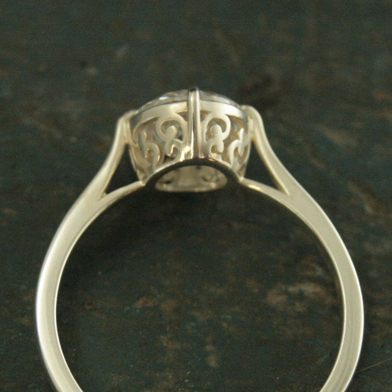 May include: A silver ring with a delicate, intricate design around the center stone. The ring is made of polished metal and has a smooth, rounded band.