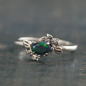 Puede incluir: Un anillo de plata con una piedra de ópalo ovalada verde oscuro engastada en un diseño floral. El anillo presenta delicados detalles de hojas y flores.
