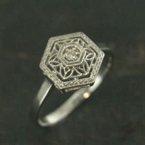 May include: A silver ring with a hexagonal setting featuring a small, clear diamond in the center. The setting is decorated with intricate filigree details.