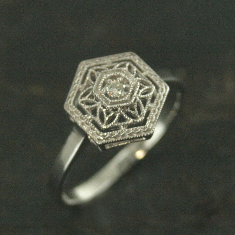 May include: A silver ring with a hexagonal setting featuring a small, clear diamond in the center. The setting is decorated with intricate filigree details.