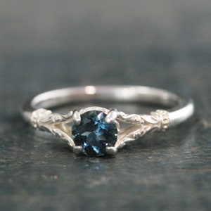 Puede incluir: Un anillo de plata con un diseño delicado y ornamentado presenta una piedra preciosa redonda de color azul oscuro.