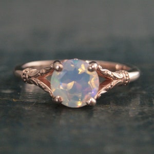 Puede incluir: Un delicado anillo de oro rosa con una piedra de ópalo blanco engastada en un diseño de inspiración vintage. El anillo presenta intrincados detalles florales en la banda.