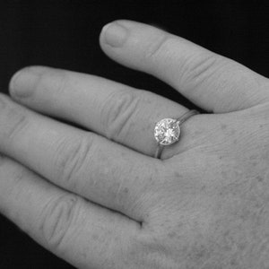 May include: A close-up of a hand wearing a silver ring with a large, round, clear diamond.