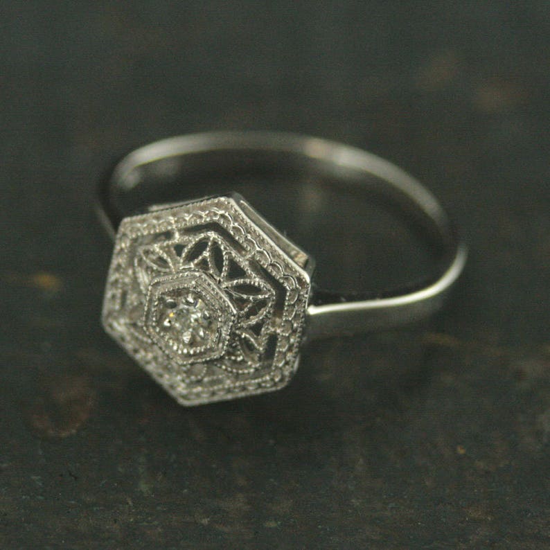 May include: A silver ring with a hexagonal setting featuring a delicate filigree design and a small, clear diamond in the center.