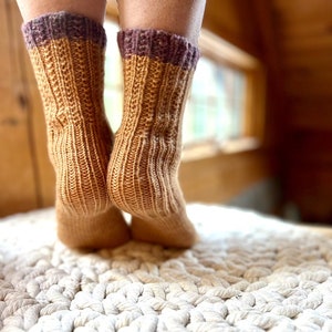 Op de afbeelding: Een paar gebreide sokken in mosterdgeel met een paarse band aan de bovenkant. De sokken staan op een wit gehaakt kleed.