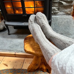 Peut inclure: Une paire de chaussettes en tricot gris avec un motif en câble, reposant sur un tabouret en bois devant une cheminée. Les chaussettes sont portées par une personne assise devant la cheminée.