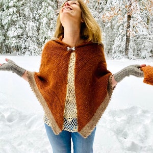 May include: A woman wearing a brown hooded poncho with a white crocheted top and blue jeans. She is standing in a snowy forest and smiling.