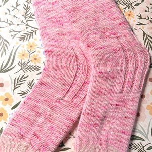 Puede incluir: Un par de calcetines tejidos a mano en tonos rosas con detalles blancos. Los calcetines presentan un puño y talón acanalados, con un patrón moteado en todo el hilo rosa. Los calcetines están hechos de un material suave y cálido.