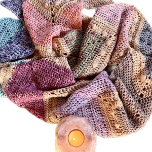 May include: A multi-colored crochet scarf featuring a textured chevron pattern with bobble stitches in shades of pink, purple, blue, and tan, positioned above a glowing, round, translucent pink salt crystal candle holder with a lit tea light.
