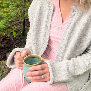 Op de afbeelding: Een persoon draagt een lichtgrijze cardigan over een roze gebloemde top en houdt een teal en gele keramische mok vast.