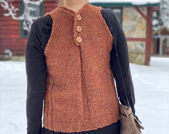 May include: A brown knitted vest with three wooden buttons down the back. The vest is worn over a black long-sleeved top and paired with olive green corduroy trousers. A brown shoulder bag is also visible.