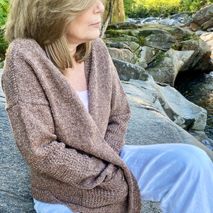 May include: A brown knit cardigan with a loose fit and a textured pattern. The cardigan is worn over a white tank top and white pants.