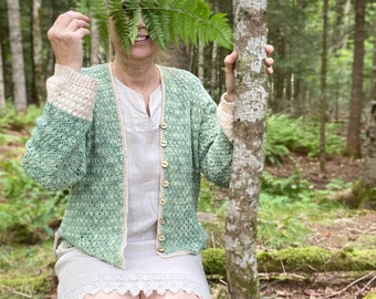 May include: A light green crocheted cardigan with beige cuffs and wooden buttons. The cardigan is worn over a light-colored dress. The image is taken outdoors in a forest setting.