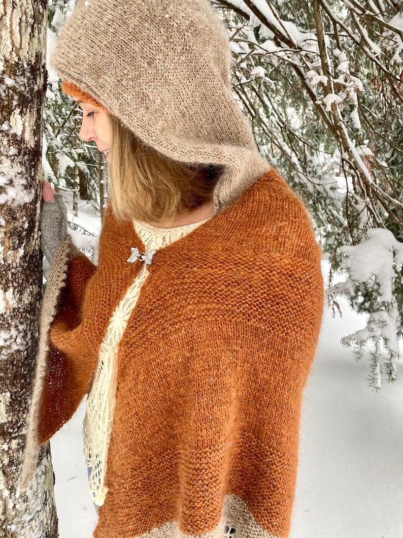 May include: A brown and beige knitted hooded shawl with a white lace trim. The shawl is worn by a person in a snowy forest setting.