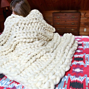 May include: A white chunky knit blanket draped over a bed with a red and black patterned bedspread.