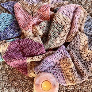 May include: A hand-crocheted shawl featuring a variegated color palette of soft pink, deep magenta, muted lavender, and sandy beige. The textile displays a repeating chevron stitch pattern with small raised bobble accents throughout the fabric.
