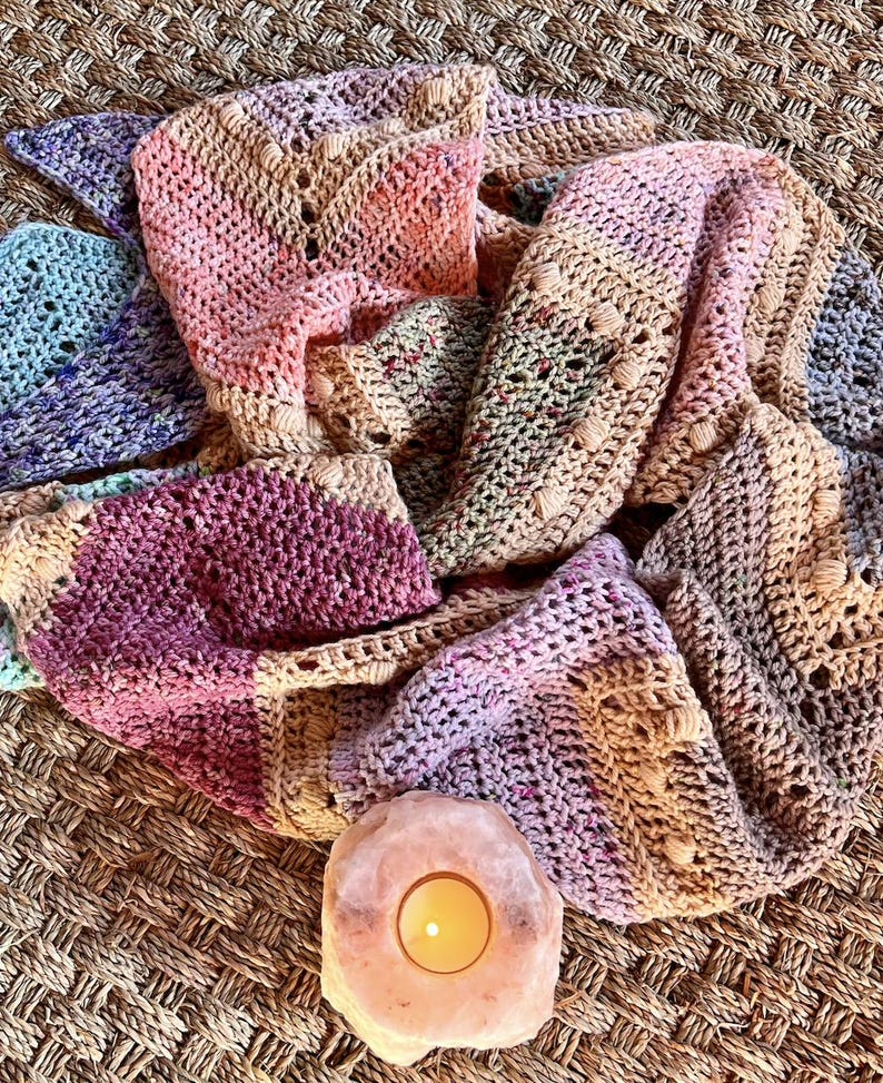 May include: A hand-crocheted shawl featuring a variegated color palette of soft pink, deep magenta, muted lavender, and sandy beige. The textile displays a repeating chevron stitch pattern with small raised bobble accents throughout the fabric.