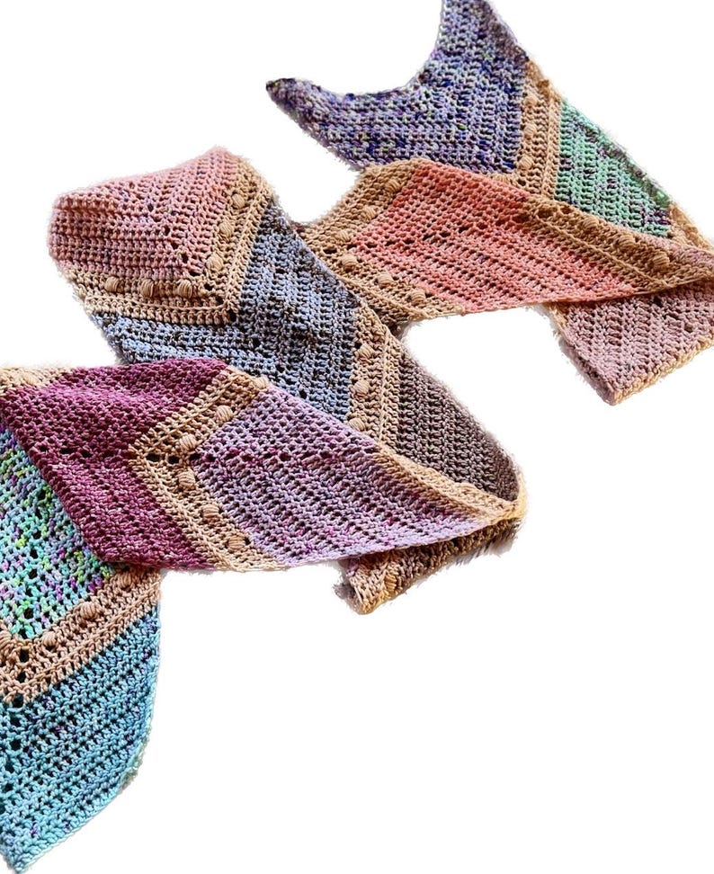 May include: A long, triangular crochet scarf featuring a multi-colored gradient pattern. The design incorporates various yarn colors including pink, blue, purple, green, and tan, with a textured stitch pattern and small bobble accents along the edges.