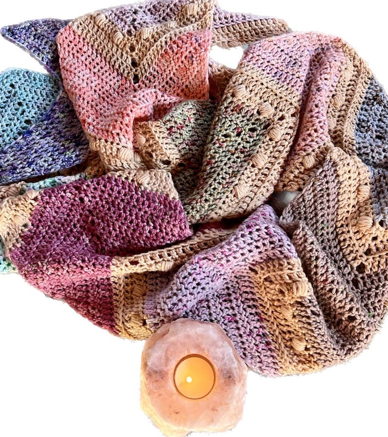 May include: A multi-colored crochet scarf featuring a textured chevron pattern with bobble stitches in shades of pink, purple, blue, and tan, positioned above a glowing, round, translucent pink salt crystal candle holder with a lit tea light.